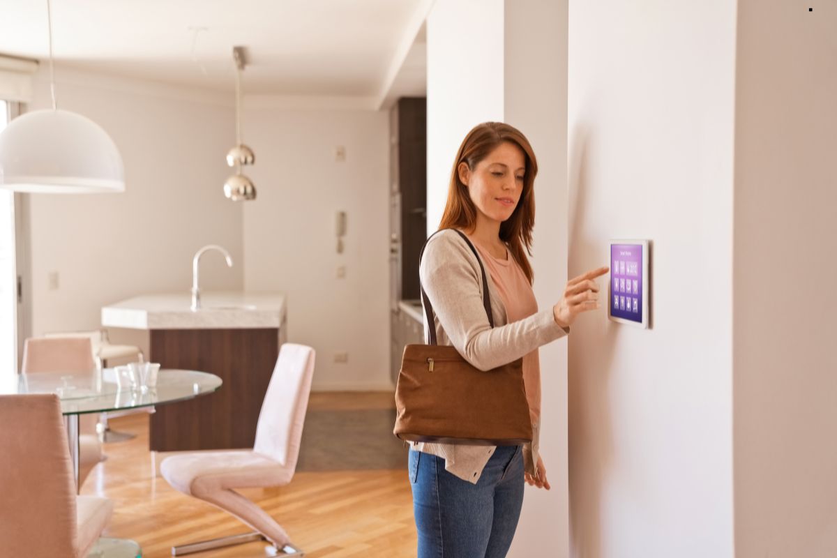 A woman stands before a wall-mounted touch screen, engaged in home security management.